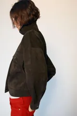 Cazadora tipo bomber de tejido efecto ante color marrón oscuro, con cuello subido, manga larga y cierre frontal con cremallera y botones ocultos. Presenta bolsillos frontales con solapa y botones metálicos.