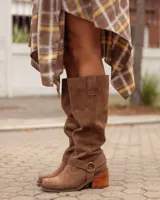 Botas de caña alta confeccionadas en cuero gamuzado color marrón. Presentan un diseño de estilo western con puntera cuadrada, taco de madera y una tira decorativa con estribo metálico desmontable en la zona del tobillo.