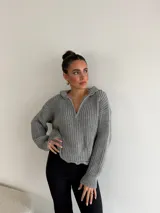 Buzo de punto grueso color gris, con cuello polo, cierre frontal parcial y capucha. Presenta un diseño de tejido acanalado con terminaciones desflecadas en el ruedo.