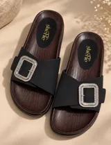 Sandalias planas color marrón con suela de aspecto amaderado y plantilla negra con logo dorado. Poseen una tira ancha de gamuza en el empeine decorada con una hebilla rectangular grande con pedrería brillante.