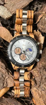 Reloj cronógrafo con caja de madera de zebrano y bisel técnico de acero negro. La esfera es plateada con subdiales de madera, indicador de fase lunar y fechador. La malla combina eslabones de acero inoxidable con madera de zebrano.