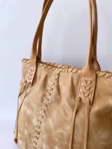 Cartera tipo tote de cuero color camel, con diseño artesanal que presenta costuras decorativas visibles en los laterales y el frente. Cuenta con doble asa de hombro y cierre superior.