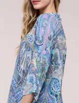 Blusa holgada de poliéster con estampado de arabescos en tonos azules, escote en V con botones frontales, mangas 3/4 y ruedo asimétrico en punta.