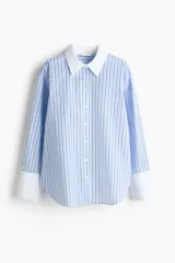 Camisa oversized de popelina de algodón con diseño a rayas verticales en tonos azul y blanco. Presenta cuello clásico y puños anchos en color blanco sólido, cierre frontal con botones, hombros caídos y bajo redondeado con aberturas laterales.