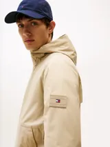 Campera beige de nailon repelente al agua y resistente al viento, con capucha fija, cierre de cremallera, dos bolsillos en la cintura, bolsillo interior con cremallera, puños elásticos con botones a presión, solapa de protección en la parte trasera y parche con el logo de Tommy Hilfiger en la manga.
