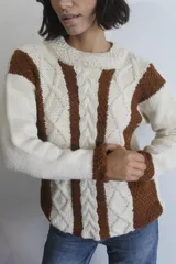 Sweater de tejido de punto artesanal en lana merino, con diseño de rayas verticales en colores crudo y marrón. Presenta un patrón de trenzas y textura en relieve, con cuello redondo y mangas largas.