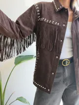 Chaqueta de efecto gamuza en color chocolate, con diseño de flecos en el pecho y mangas. Presenta detalles de tachas metálicas plateadas a lo largo de los hombros, solapas y bolsillos. Cuenta con cierre frontal de botones y forro interior.