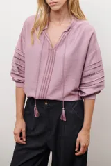 Camisa de color crudo con diseño romántico, cuello redondo con cordón ajustable terminado en borlas, y mangas tres cuartos abullonadas. Presenta detalles de bordados calados verticales en el frente y horizontales en las mangas.