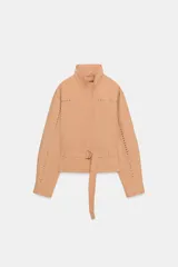Chaqueta de cuello subido y manga larga, color beige. Presenta detalles de tachas metálicas a lo largo de las mangas y en la parte superior delantera. Incluye un cinturón del mismo tejido con hebilla metálica y cierre frontal con cremallera y botones a presión ocultos.