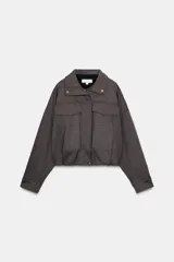 Chaqueta estilo bomber de color gris oscuro, con cuello subido, manga larga y corte holgado. Presenta bolsillos delanteros con solapa, bajo con pliegues y cierre frontal con cremallera oculta y botones a presión.