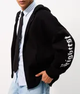 Campera con capucha de felpa negra, con cierre frontal, bolsillos canguro y estampa tipográfica gótica en color blanco en las mangas.