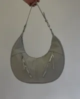 Cartera mediana tipo hobo de cuero sintético color gris, con diseño semicircular, dos bolsillos externos frontales con cierre y correa de hombro ajustable.