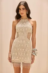 Vestido corto de crochet color beige con escote halter y espalda nadadora. Presenta inserciones de malla transparente en la cintura, bordado de perlas y cristales, y un ruedo con flecos largos de cuentas.