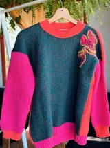 Sweater de punto con diseño color block, presentando un frente en tono verde oscuro melange con un bordado floral en el hombro, y mangas en color fucsia vibrante. Posee cuello redondo y puños acanalados.