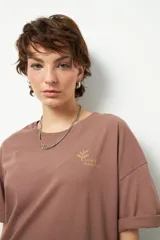 Remera de algodón color marrón topo, corte oversize y cuello redondo. Presenta un pequeño estampado frontal con un logo de hoja y texto "LOVE EARTH" en color dorado.