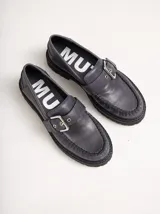 Mocasines de cuero color gris oscuro con diseño de costura en el empeine, ajuste mediante tira con hebilla metálica y suela track con taco de 4 cm.