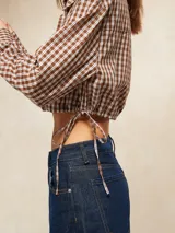 Camisa de manga larga, cuello camisero y botones frontales, con estampado a cuadros marrones y blancos. El ruedo inferior es ajustable con cordones laterales, creando un efecto *cropped* y fruncido.