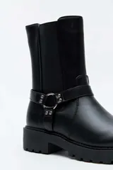 Bota alta color negro, con suela track y taco cuadrado. Presenta una tira con tachas y una argolla metálica decorativa alrededor del tobillo, además de inserciones elásticas laterales.