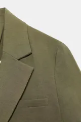 Blazer corto color verde oliva, de cuello solapa y manga corta. Tiene un bolsillo de vivo en el pecho y cierre frontal con un botón.