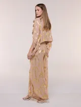 Pantalón palazzo confeccionado en gasa transparente color rosa con diseño estampado metalizado dorado. Presenta cintura elástica con cordón ajustable con borlas, piernas anchas y diseño cruzado al frente con aberturas laterales y detalles de puntillas.