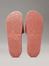 Ojotas tipo slide de color rosado, con base de EVA y correa ancha de lona de algodón reciclado. La correa presenta el logo de Calvin Klein Jeans bordado en blanco.