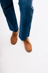 Mocasín de estilo relajado confeccionado en cuero gamuzado color tostado. Presenta detalles fruncidos en la puntera, cordones decorativos en el empeine y el logo de la marca grabado en el lateral.