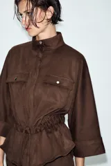 Camisa de efecto ante en color marrón, con cuello subido, mangas tres cuartos con vuelta y cierre frontal con cremallera. Presenta falsos bolsillos con solapa en el pecho y cintura ajustable con cordones del mismo tejido.