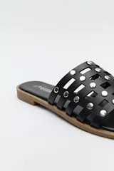 Sandalia baja negra, tipo mule o chinela, con capellada de tiras entrelazadas y apliques metálicos redondos (tachas). La suela es plana y de color marrón claro.