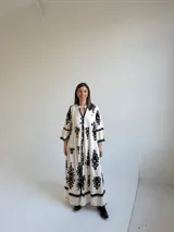 Vestido largo color marfil con estampado ornamental en color negro, escote en V con botones frontales, mangas largas amplias y diseño escalonado en la falda.