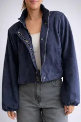 Chaqueta corta de efecto gamuza con cierre frontal y botones. Presenta mangas tipo globo con puños elásticos y detalle de aros metálicos decorativos en la solapa. Cuenta con ajuste lateral en la cintura y forro interior.