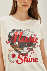 Remera de algodón color crema con estampa frontal de una bola de espejos en forma de corazón y texto en tonos rojos y negros.