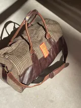 Bolso de viaje tipo duffel confeccionado en una combinación de cuero genuino marrón y tela texturizada a rayas. Cuenta con doble asa de mano, correa de hombro ajustable y desmontable, cierre superior y bolsillo interno.