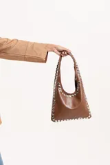 Cartera de mano tipo hobo confeccionada en eco-cuero color marrón, con diseño estructurado y borde decorado con tachas metálicas plateadas.