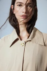 Trench midi de color camel, confeccionado en una mezcla de algodón y nylon con propiedades repelentes al agua. Presenta un diseño clásico con cuello camisero, cierre frontal de botones, bolsillos laterales y tajos en los costados.