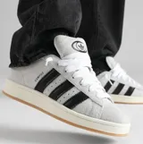 Championes Adidas Campus 00S de gamuza color gris claro con las tres tiras características de la marca en negro. Suela de goma color hueso con base marrón.