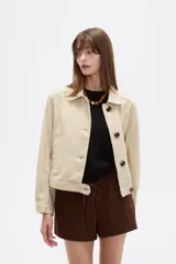 Chaqueta corta de algodón color beige, con cuello camisero, bolsillos laterales y cierre frontal con botones oscuros.
