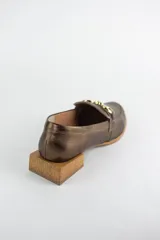Mocasines de cuero en tono bronce metalizado, con detalle de aplique metálico dorado en el empeine y taco cuadrado bajo de madera.