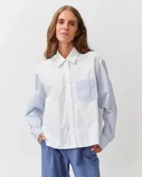 Camisa de algodón con diseño combinado. El cuerpo es de color blanco liso, mientras que las mangas largas y el bolsillo frontal aplicado presentan un patrón de rayas finas celestes y blancas. Cuenta con cuello clásico y cierre frontal de botones.