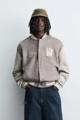 Cazadora estilo varsity o bomber de hombre, color gris oscuro con efecto ante, mangas en tono gris claro y puños, cuello y bajo con rib elástico a rayas blancas y grises. Presenta un parche con la letra 'R' en el pecho derecho y un bordado con texto en el pecho izquierdo.