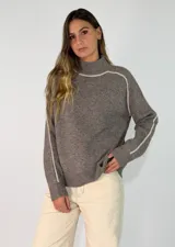 Sweater de punto color marrón topo con cuello alto y costuras decorativas en color crema que recorren los hombros y las mangas. Presenta un diseño de corte holgado con puños y ruedo acanalados.