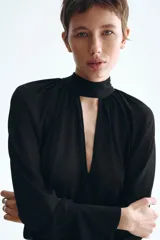 Blusa oversized negra de tejido transparente, con cuello fular, escote profundo y mangas largas con puños anchos.