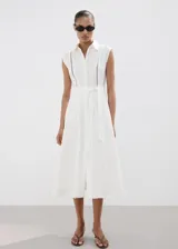 Vestido midi camisero de lino color blanco, sin mangas, con cuello solapa, cierre frontal de botones y cinturón ajustable del mismo género. Cuenta con detalles de costura calada en el frente.