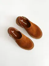 Zueco de cuero gamuzado color óxido, con tira trasera ajustable mediante hebilla metálica. Presenta una suela de plataforma con textura acanalada y detalle de textura reptil en la tira posterior.