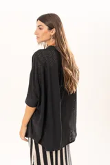 Camisa negra de corte oversize con diseño de encaje calado, escote en V, cierre frontal con botones y mangas tres cuartos. Presenta un diseño asimétrico con la parte posterior ligeramente más larga.