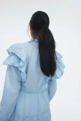 Vestido corto de algodón celeste, con cuello alto con volados y abertura frontal con botones. Presenta mangas largas, elástico en la cintura y falda escalonada con detalles de encaje y volados en los bordes.