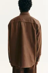 Camisa de corte oversized en denim de algodón rígido revestido, color marrón oscuro. Presenta cuello inglés, tapeta clásica con botones de metal y canesú en la espalda. Tiene hombros caídos, mangas largas con puños abotonados y bajo redondeado.