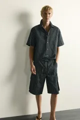 Bermuda cargo relaxed fit confeccionada en denim de mezcla de algodón y lino, con diseño de rayas verticales grises y negras. Cuenta con bolsillos laterales tipo cargo con solapa y cierre frontal con cremallera y botón.