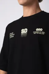 Remera de corte oversize color negro, confeccionada en algodón premium. Presenta un diseño gráfico minimalista estampado en el frente con tipografía y logotipos en color crema.
