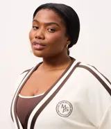 Cardigan deportivo de corte oversized, color blanco con detalles en marrón oscuro. Presenta escote en V, cierre frontal con botones a presión, mangas largas con franjas laterales y puños acanalados. Incluye un parche circular con letras en el pecho.