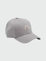 Gorra de béisbol de color gris liso, con visera curva y el logo circular de Moncler bordado en el frente.